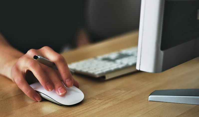 Free A hand using a wireless mouse at a modern desk setup with a computer and keyboard. Stock Photo